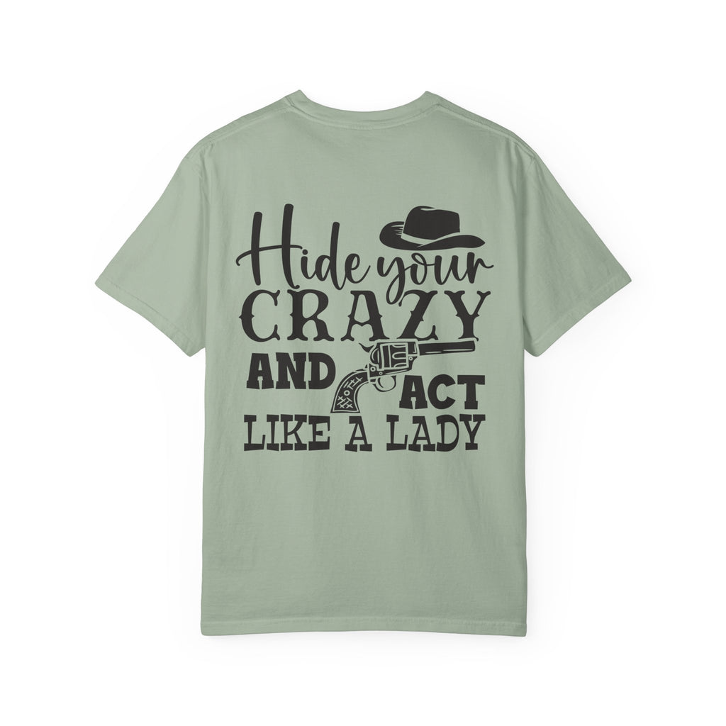 A sage green short-sleeve t-shirt with a black graphic print on the back featuring the text 'Hide your CRAZY AND ACT LIKE A LADY' along with illustrations of a cowboy hat and a revolver, shown against a plain white background.