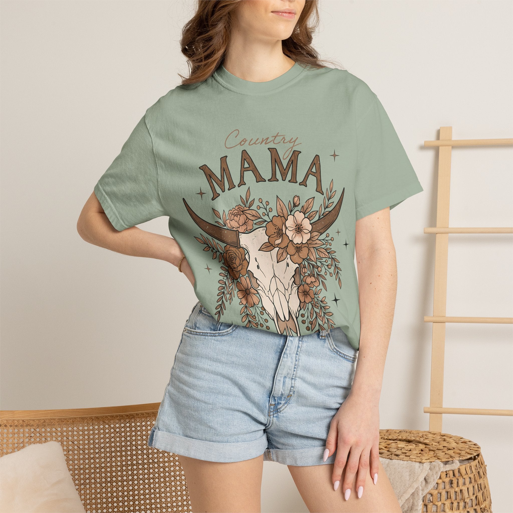 A woman wearing a sage green graphic t-shirt with a 'Country Mama' cow skull and floral design, paired with light-wash denim shorts in a cozy indoor setting.