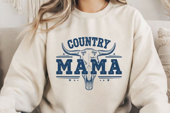 A person wearing a cream sweatshirt with a dark blue graphic that reads 'COUNTRY MAMA' above a longhorn skull design, shown in a cozy indoor setting.